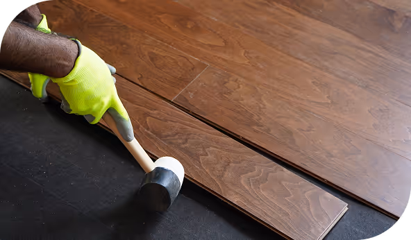 Close-up of a person installing wooden flooring with a rubber mallet and wearing safety gloves, demonstrating a step in the flooring installation process.