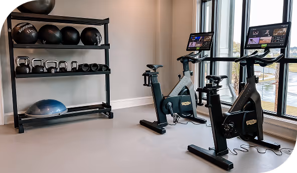 Modern gym setup with exercise bikes, kettlebells, medicine balls, and a balance trainer near large windows, highlighting versatile rubber flooring applications.