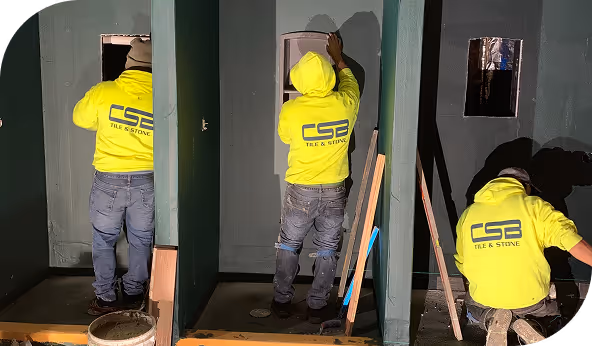 Workers installing tile and stone flooring in a commercial space, wearing C.S.B. Tile & Stone branded jackets, focusing on construction and renovation.