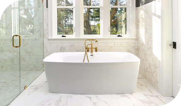 Luxurious bathroom with a freestanding white bathtub, marble tile backsplash, and gold fixtures in a bright, window-lit space.
