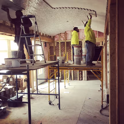 Construction workers installing ceiling panels on scaffolding inside a building under renovation, with exposed brick walls and insulation visible.