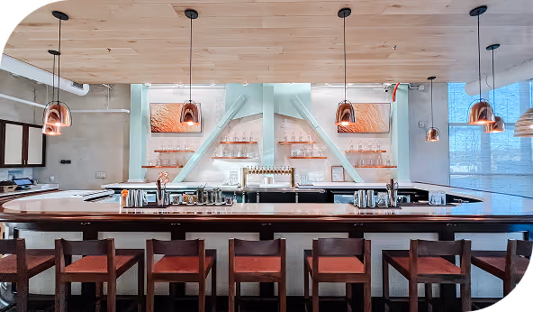 Modern bar interior with wooden ceiling, copper pendant lights, and a sleek countertop in a commercial setting.