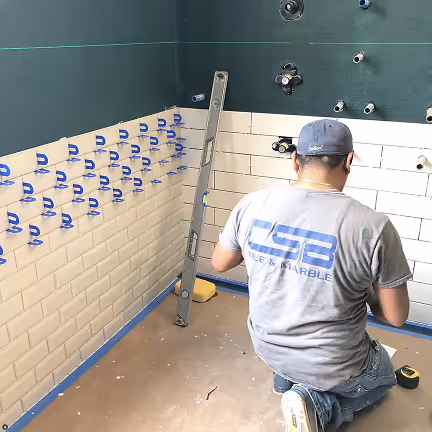 Worker installing white subway tiles on a bathroom wall with blue spacers, using a level for alignment. CSB Tile & Marble logo visible on shirt. ADA compliant.