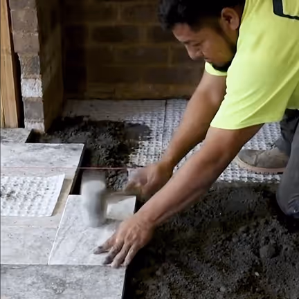 Worker installing brown floor tiles with a rubber mallet on a construction site.