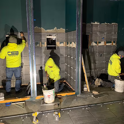 Three workers in neon CSB uniforms installing wall tiles inside commercial building under construction