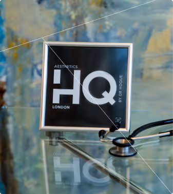 Square black sign with white text reading 'Aesthetics HQ London by Dr Roque' on a glass table next to a stethoscope.