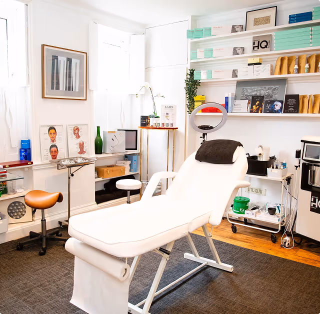 Bright and clean treatment room with a white reclining chair, medical stool, shelves stocked with skincare products, and anatomical face charts on the wall.