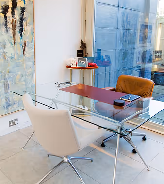 Modern glass office desk with white and brown chairs, a red desk pad, notebook, and small table with decorative items by a large window.