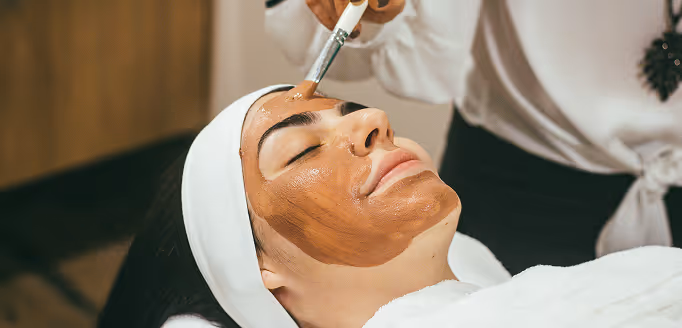 Woman receiving a facial treatment with a brown mask being applied using a brush while lying down with eyes closed and wearing a white headband and towel.