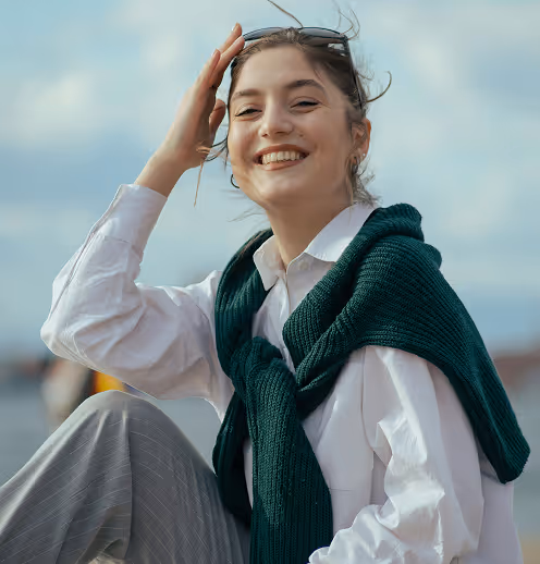 woman smiling outdoors wearing a white shirt with a dark green sweater draped over her shoulders and holding sunglasses on her head