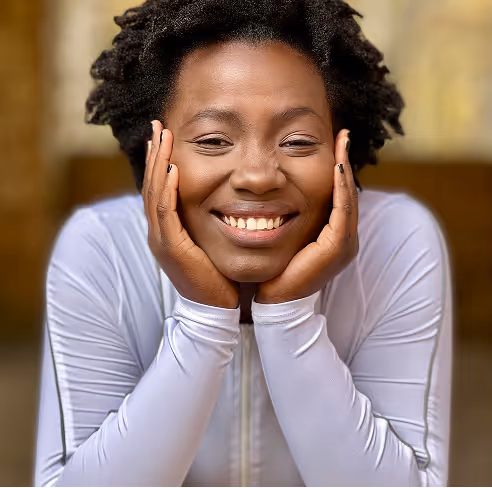 woman with short curly hair smiling warmly, resting her face in her hands, wearing a fitted white long-sleeve top indoors