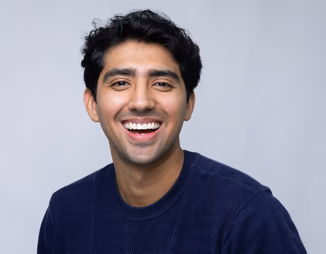 portrait of a young man with dark hair smiling broadly, wearing a navy blue sweater against a light grey background