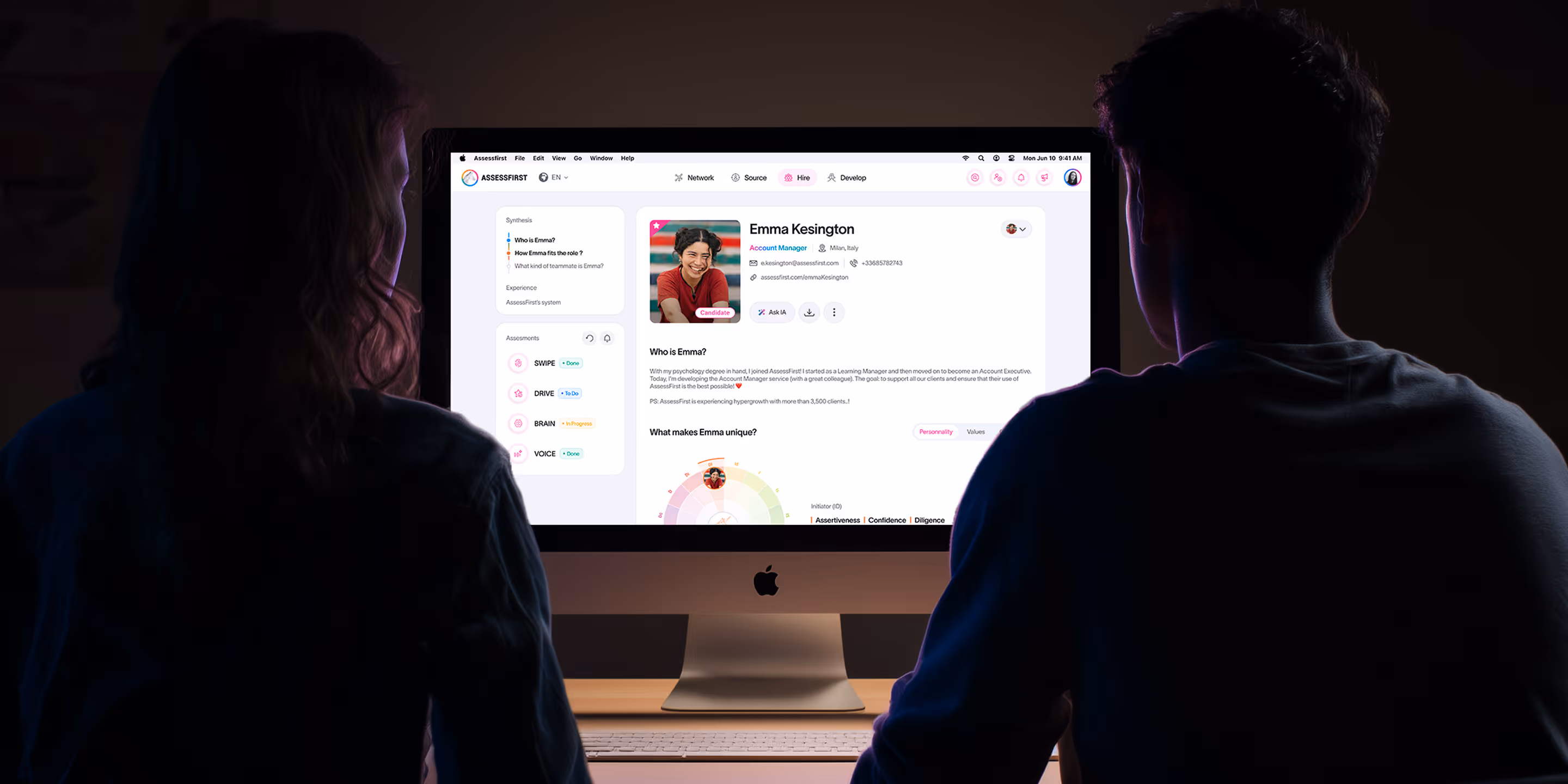 Two people sitting in front of a desktop computer in a dim room, viewing a candidate profile and assessment dashboard.
