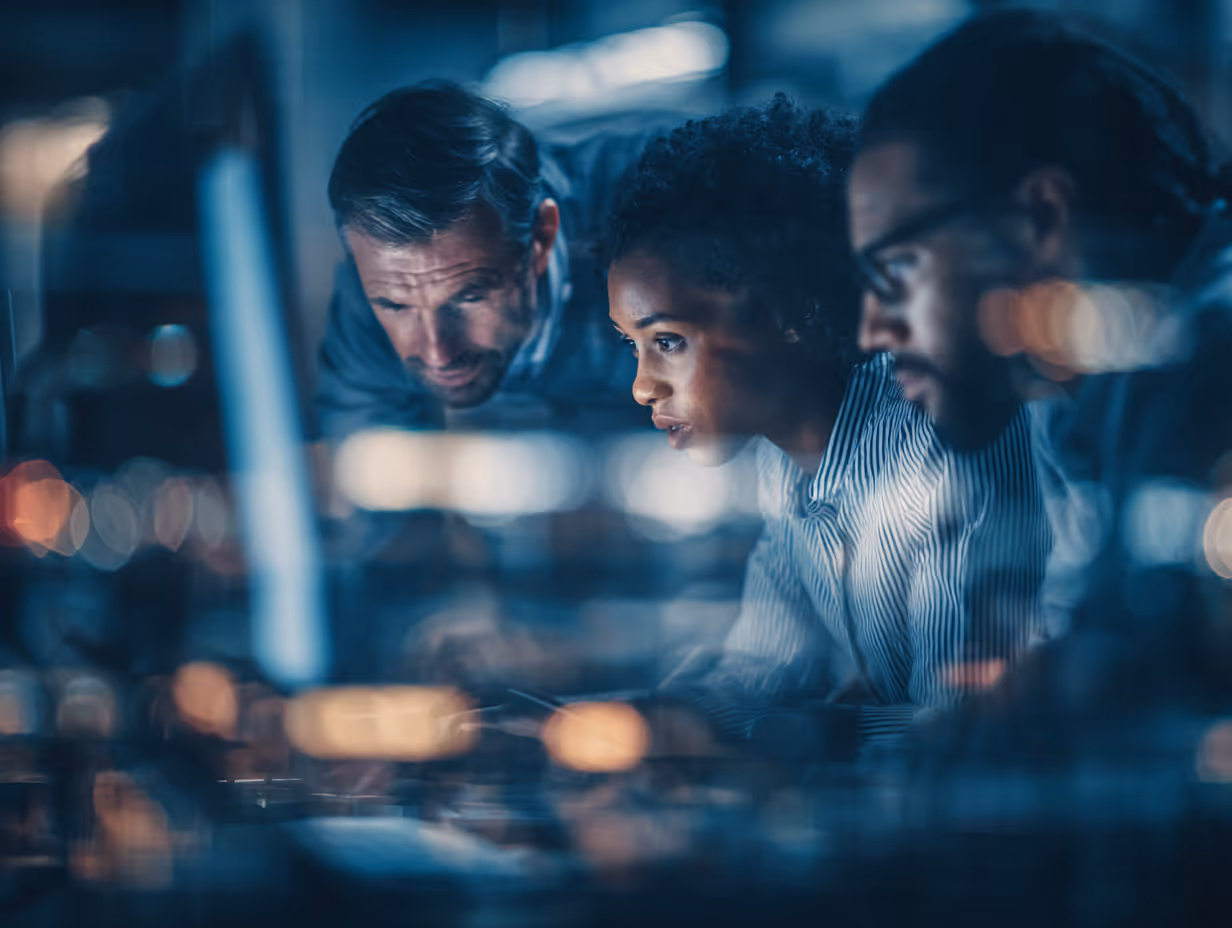 Three professionals focused on a computer screen, working late in an office with blurred lights in the foreground.