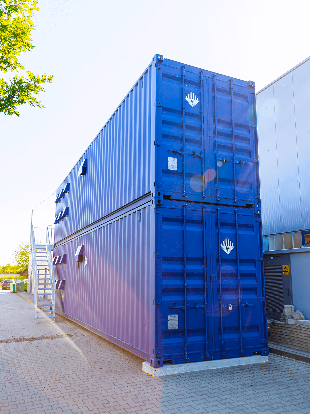 Zwei übereinander gestapelte blaue Container auf einem gepflasterten Platz, umgeben von Gebäuden und Bäumen.