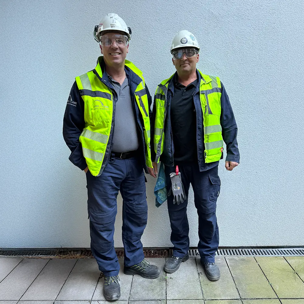 Zwei Männer in Warnwesten, Schutzhelmen und Schutzbrillen stehen vor einer weißen Wand.