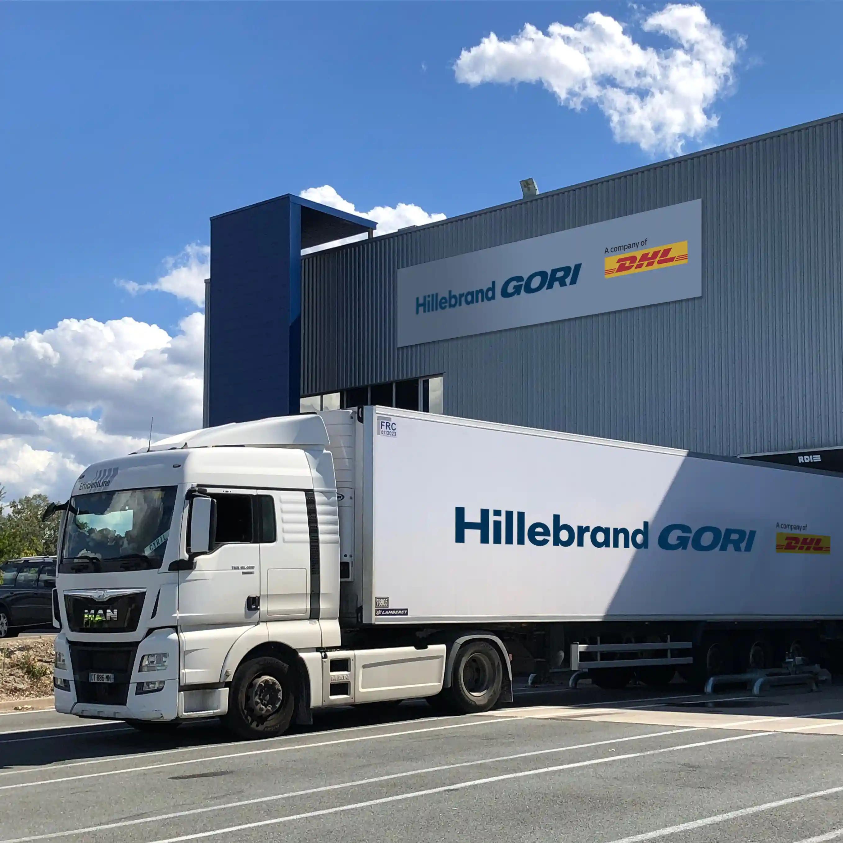 Hillebrand GORI freight truck parked at a warehouse facility with company signage visible on the building. The trailer features logos for Hillebrand GORI and DHL.