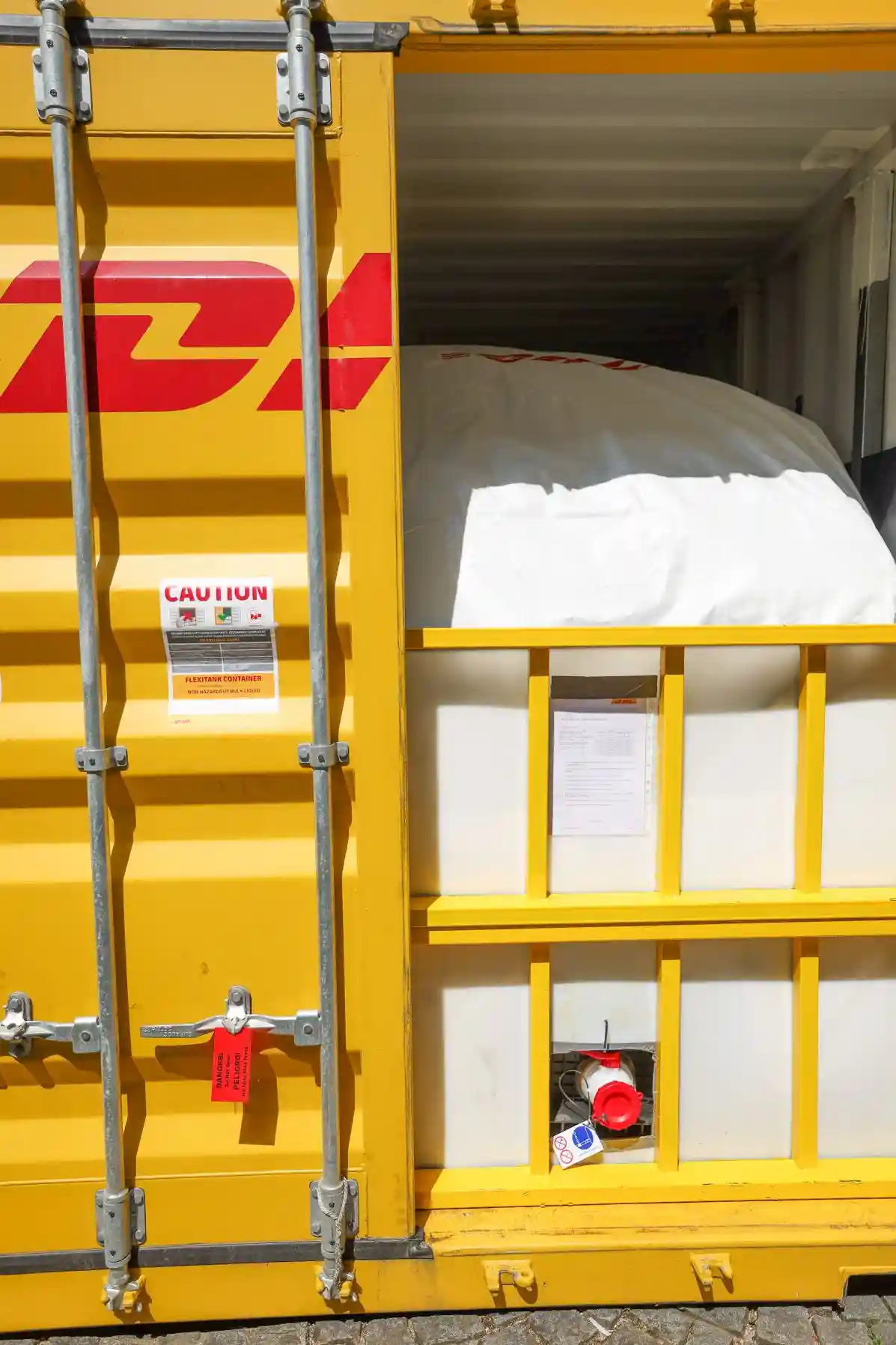 Open yellow DHL shipping container showing a flexitank filled with liquid cargo, secured inside with a bulkhead barrier and visible valve outlet.