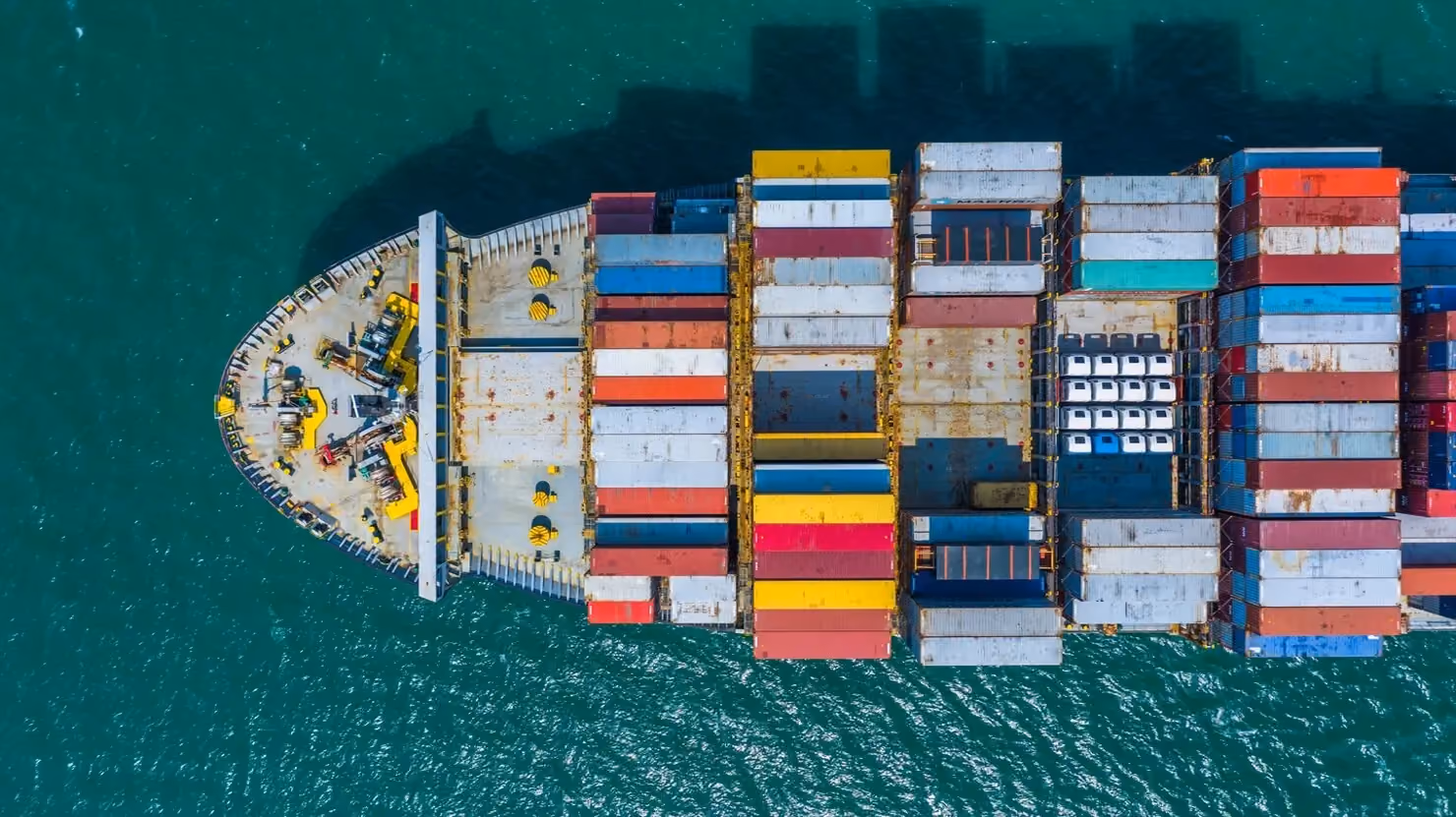Cargo ship carrying stacked containers being guided by tugboats into a busy commercial port.