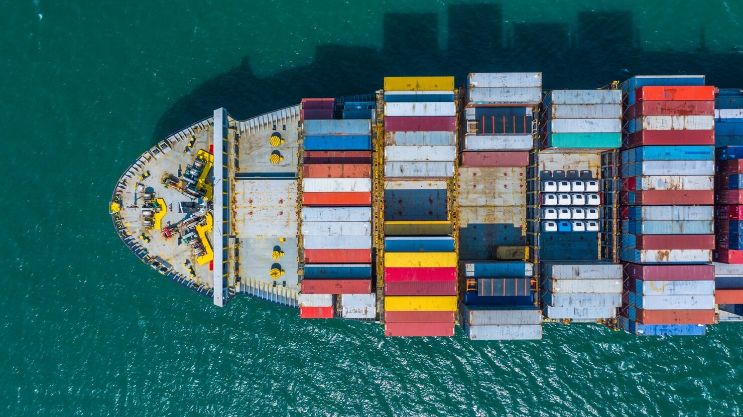 Cargo ship carrying stacked containers being guided by tugboats into a busy commercial port.