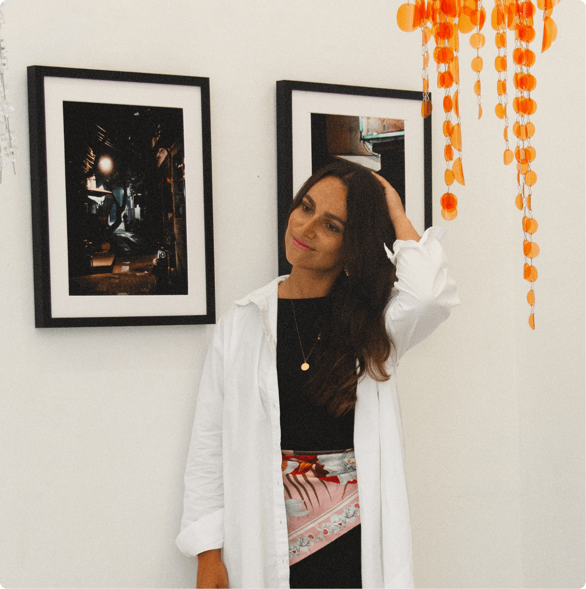 Woman with long dark hair wearing a black top, white open shirt, and floral scarf stands against a white wall with framed photos and hanging orange decorations.