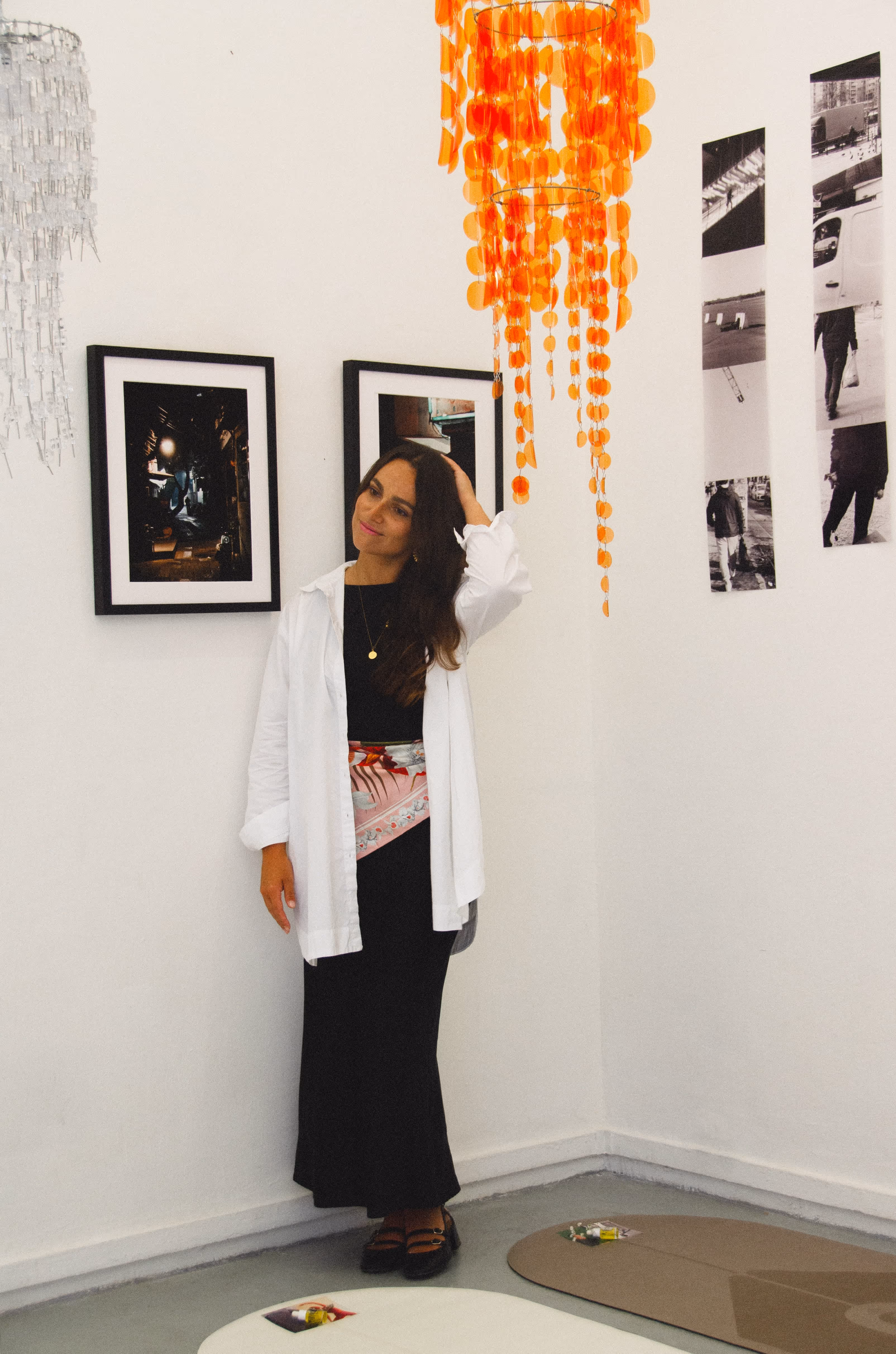 Woman in a long black dress and white shirt standing in a gallery with framed artwork and orange and clear hanging decorations.