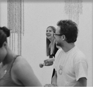 Black and white photo of three people smiling and interacting indoors with hanging decorations in the background.