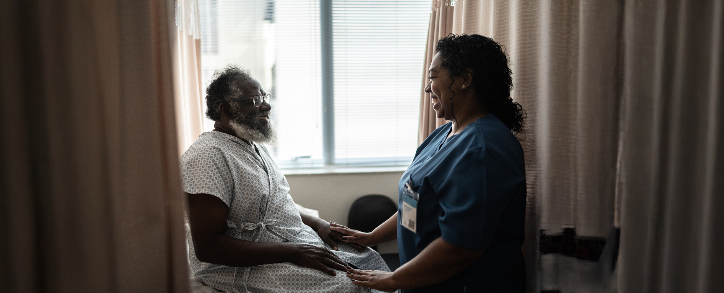 Nurse smiling and holding hands with an elderly male patient wearing a hospital gown, seated by a window with blinds.