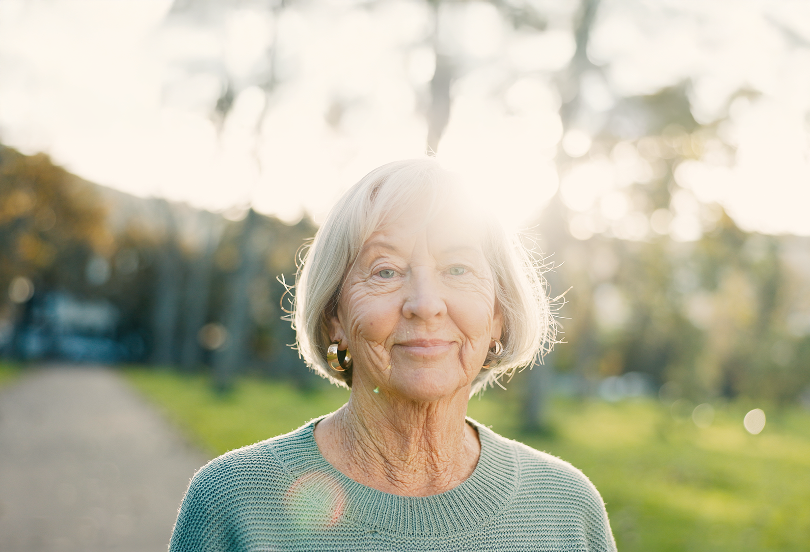 Smiling elderly woman with short gray hair wearing a green sweater outdoors with sunlight behind her.
