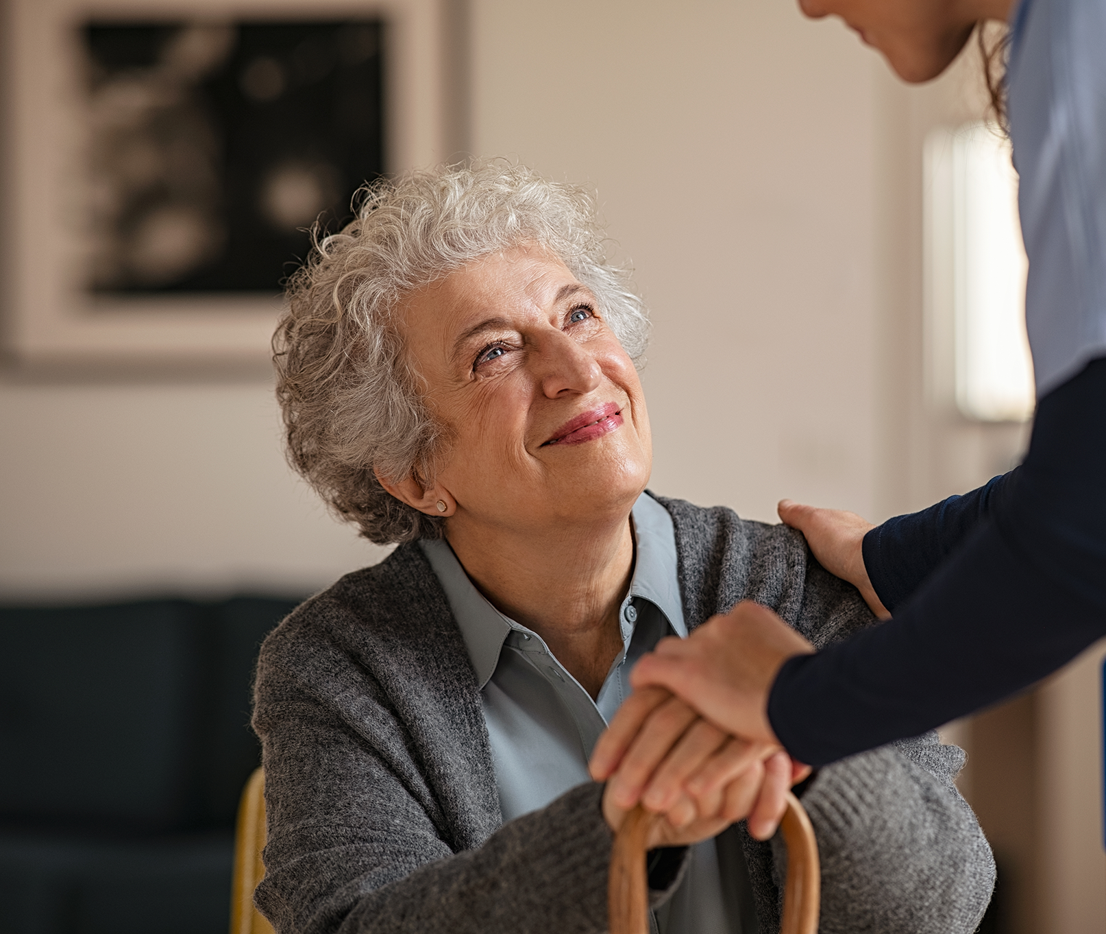 Smiling elderly woman with gray curly hair holding a cane and looking up at a caregiver who is holding her hands and shoulder.