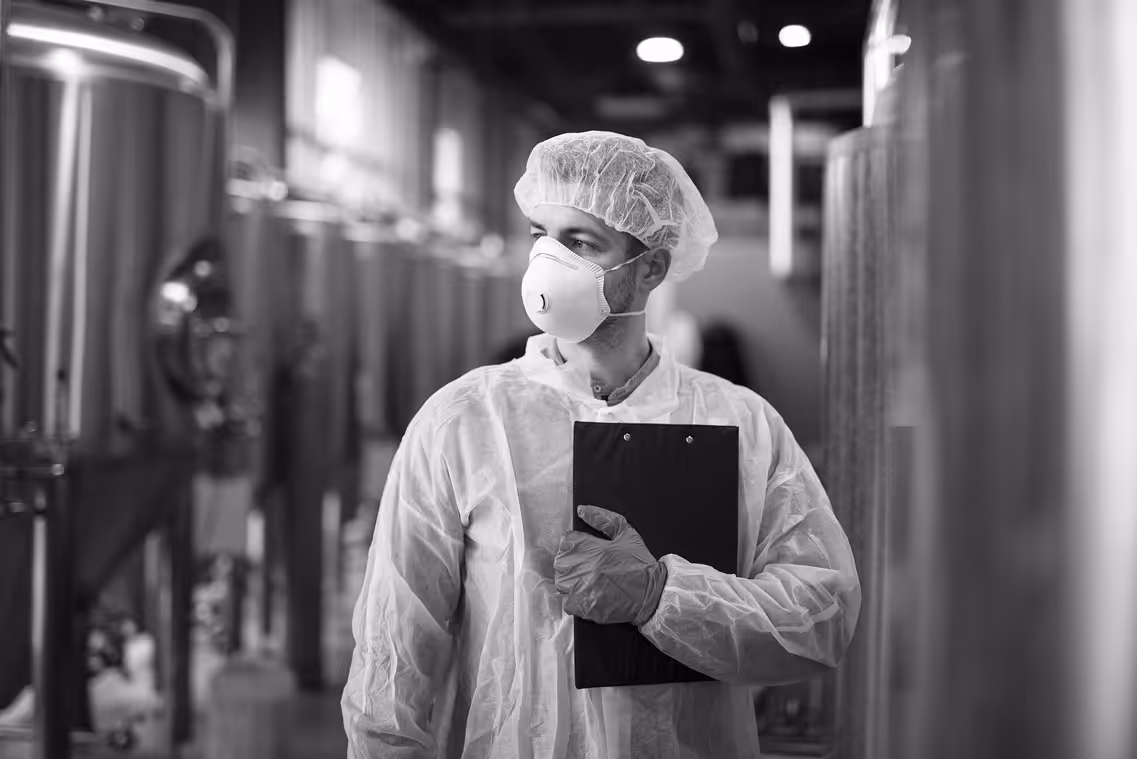 Worker in protective suit, hairnet, gloves, and mask holding a clipboard in a factory or industrial setting.