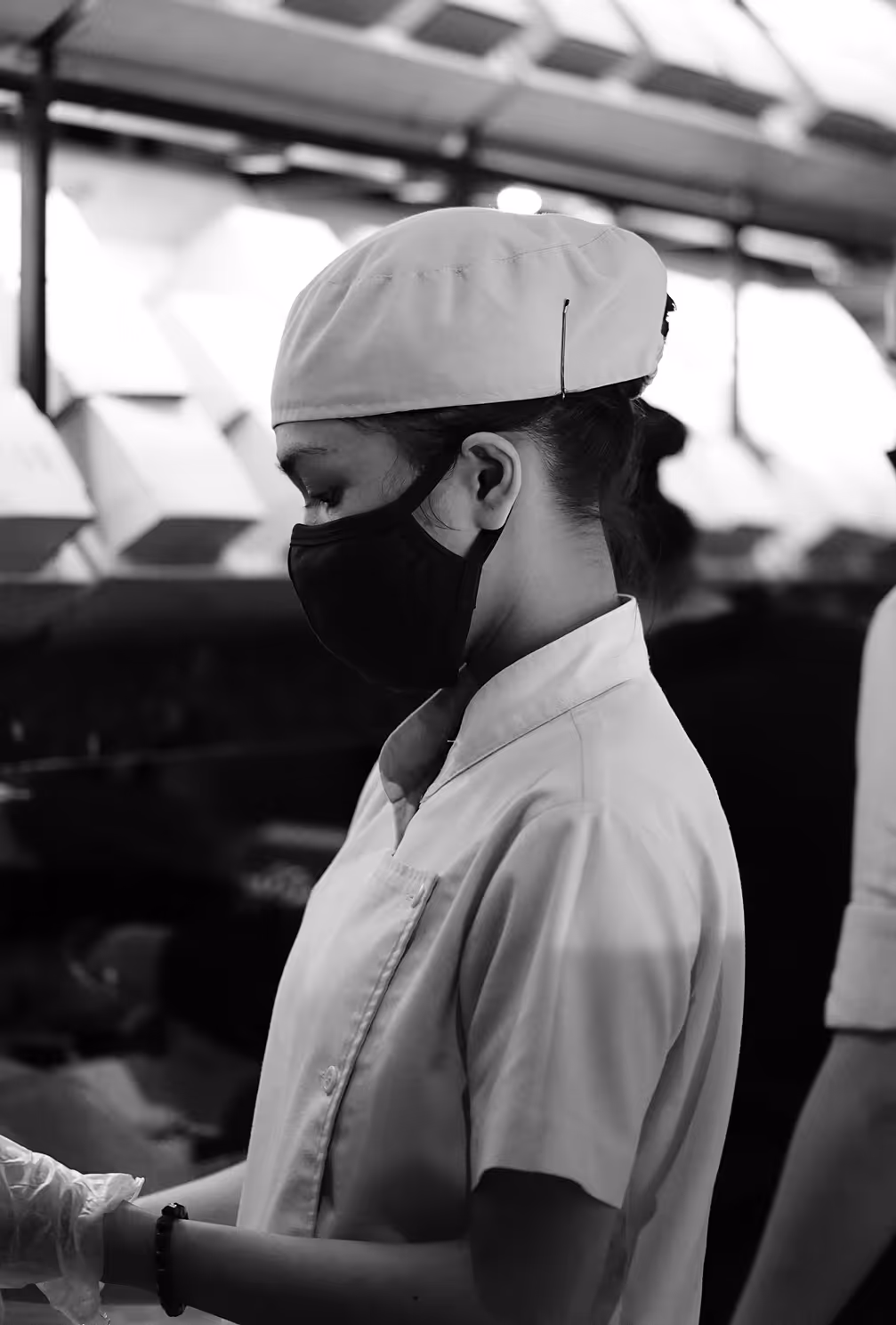 Side profile of a woman wearing a chef's hat, face mask, and gloves, working in a kitchen.