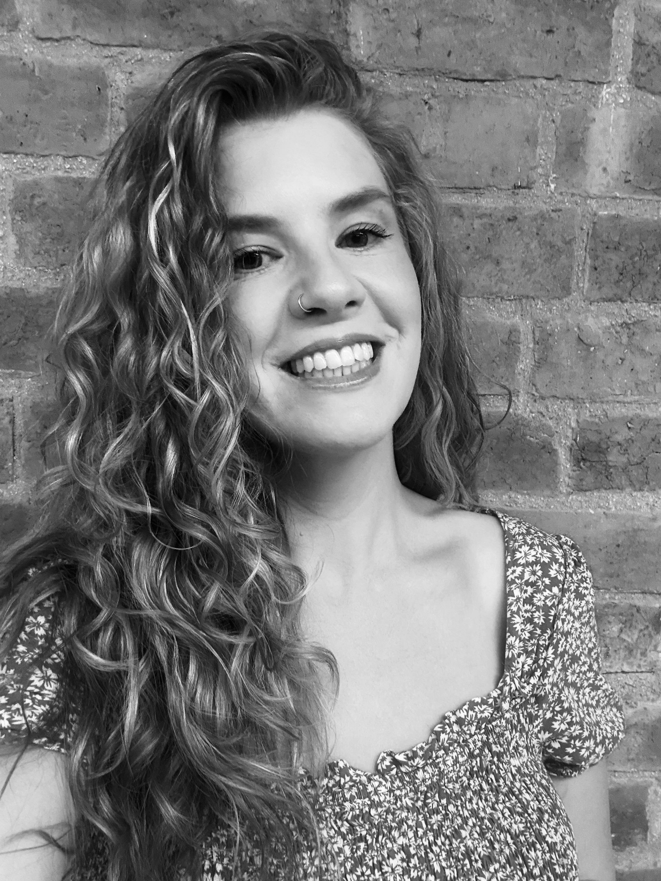 Smiling woman with short blonde hair wearing a patterned jacket and a necklace, standing in front of a brick wall.
