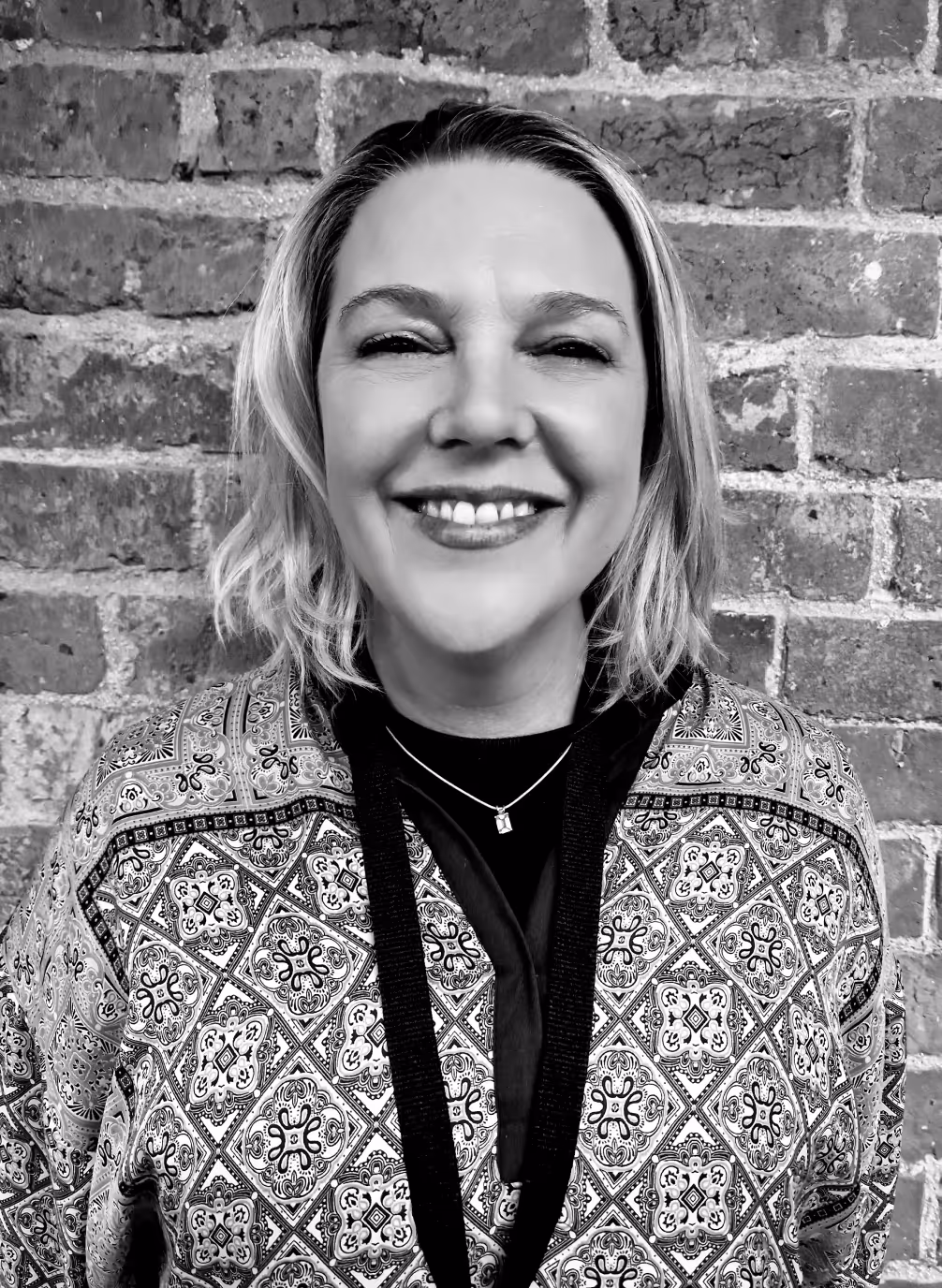 Smiling woman with short blonde hair wearing a patterned jacket and a necklace, standing in front of a brick wall.