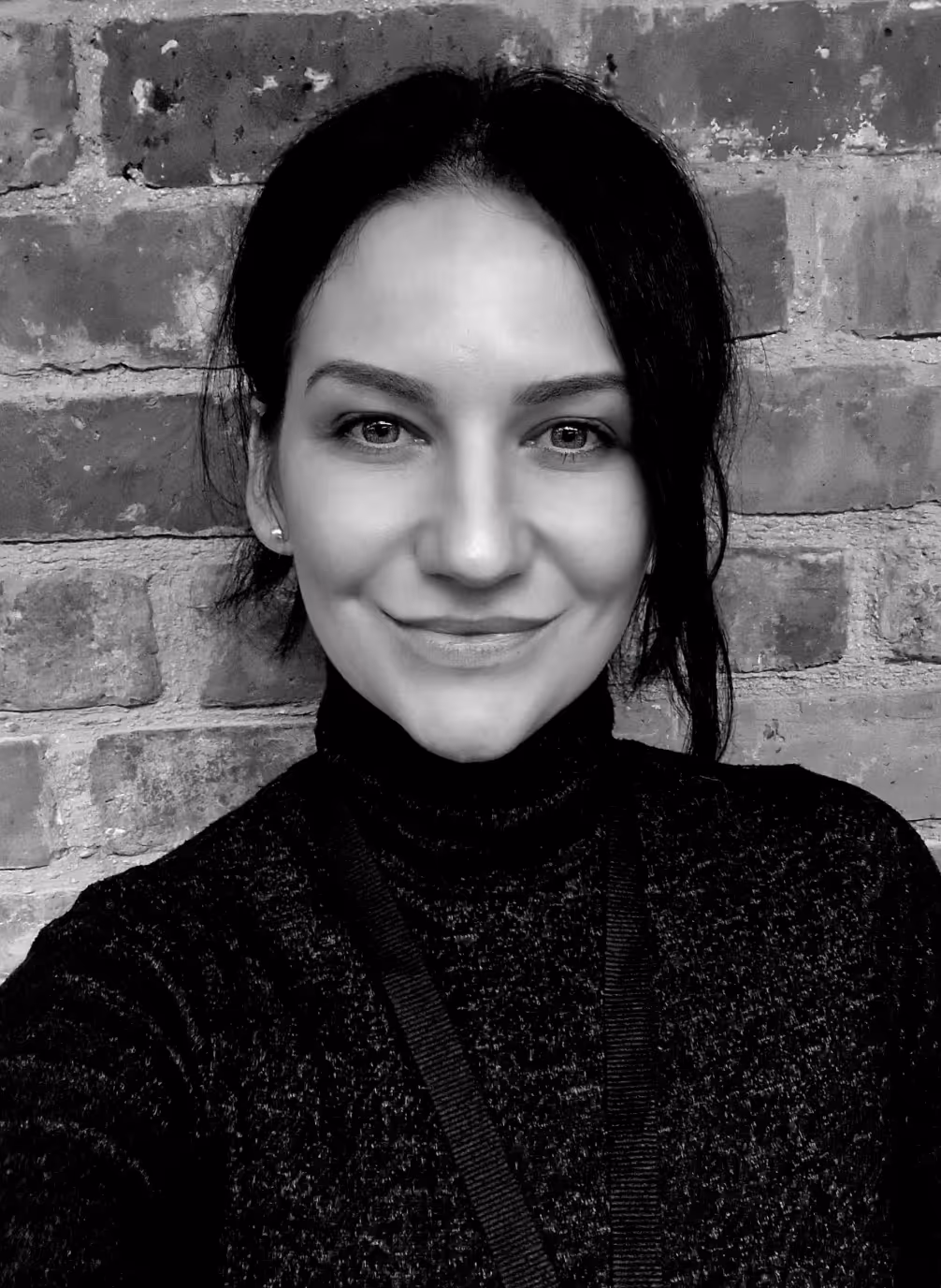 Black and white portrait of a smiling woman with dark hair wearing a turtleneck sweater against a brick wall.