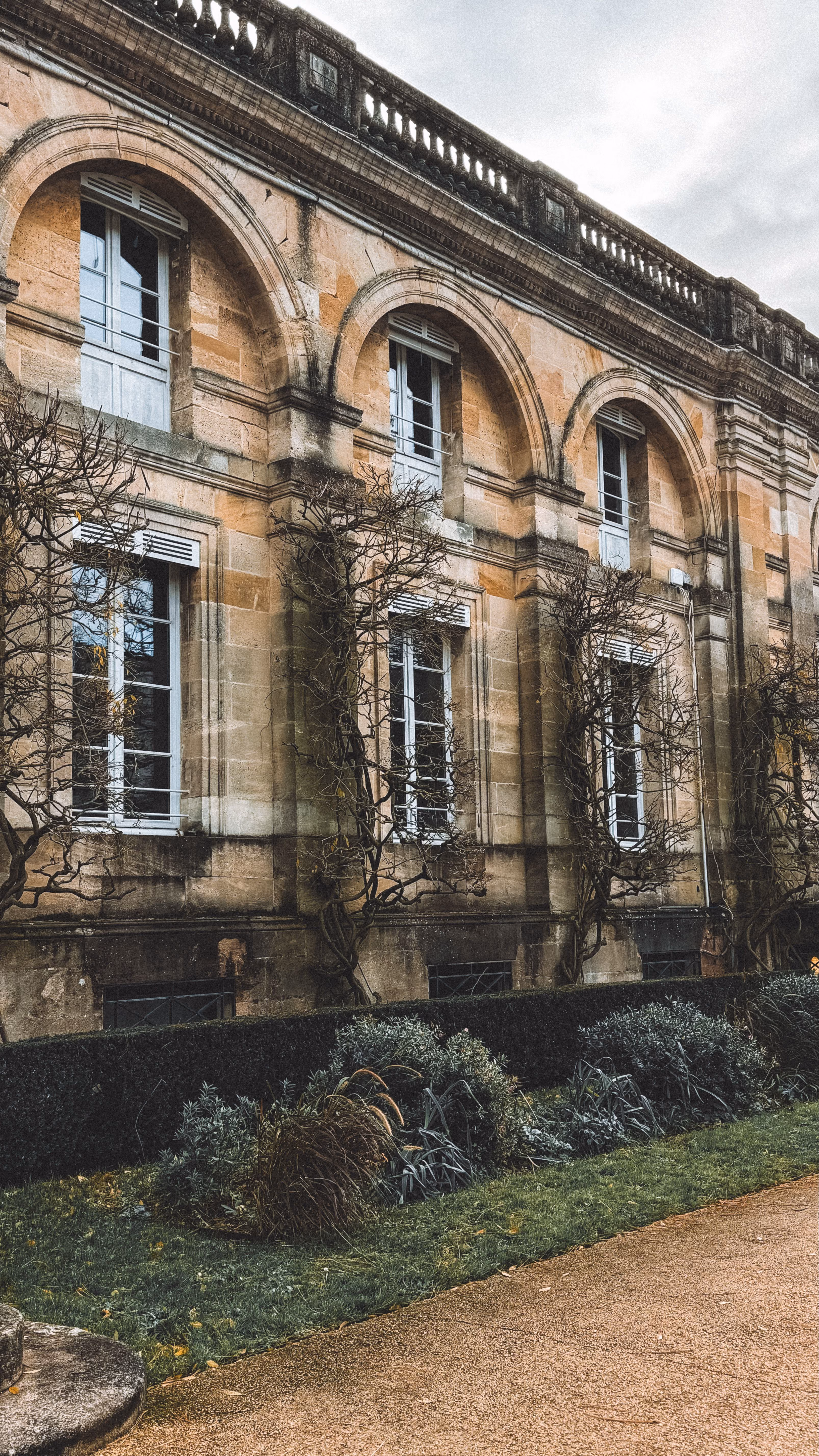 Façade en pierre beige avec grandes fenêtres à arcades et haies taillées devant.