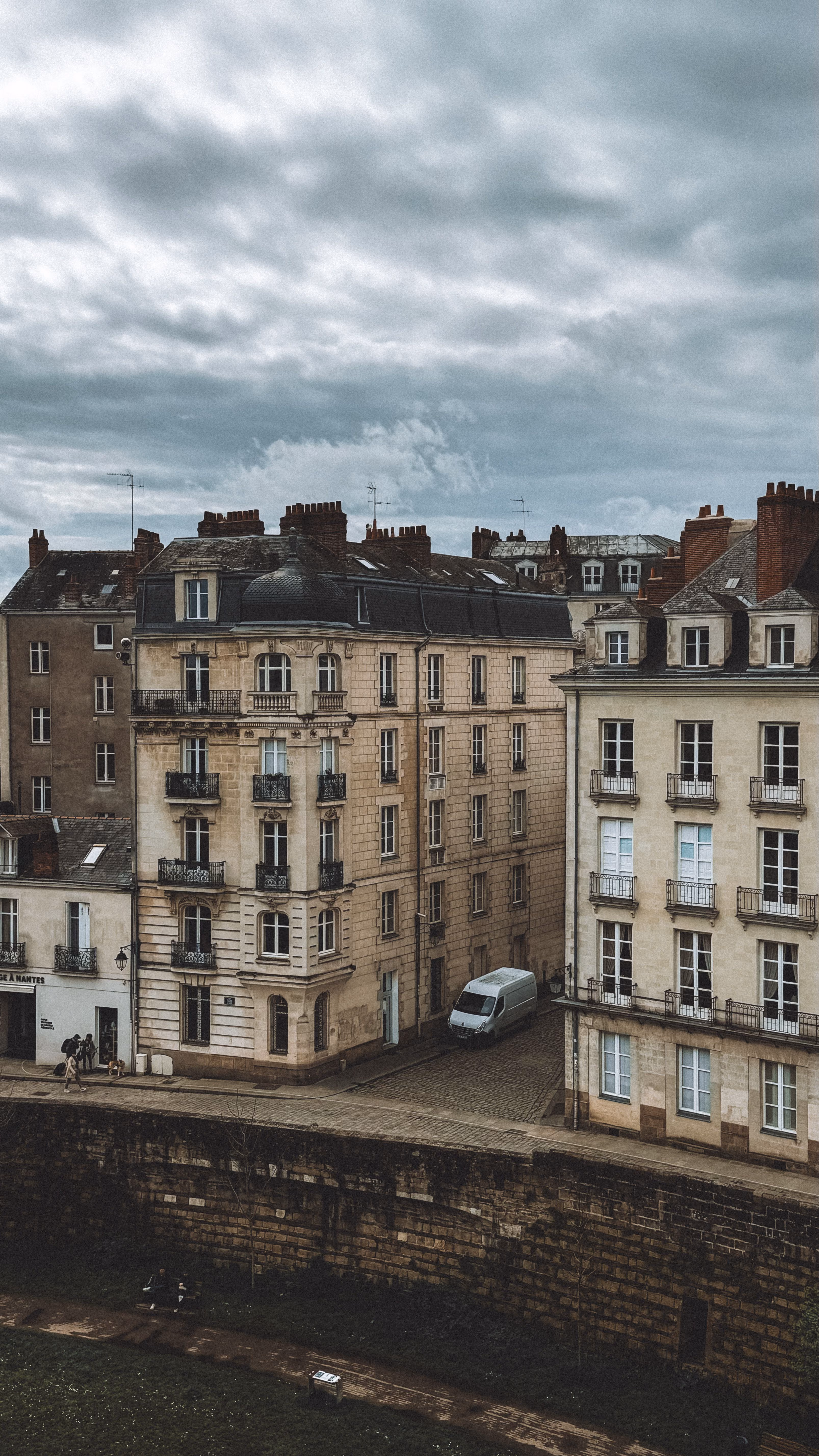 Bâtiments anciens à plusieurs étages sous un ciel nuageux avec un van blanc stationné dans une ruelle.