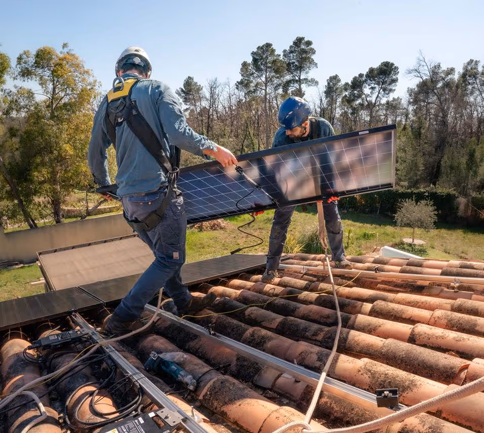 Deux installateurs portant un casque installent un panneau solaire sur un toit en tuiles sous un ciel clair.