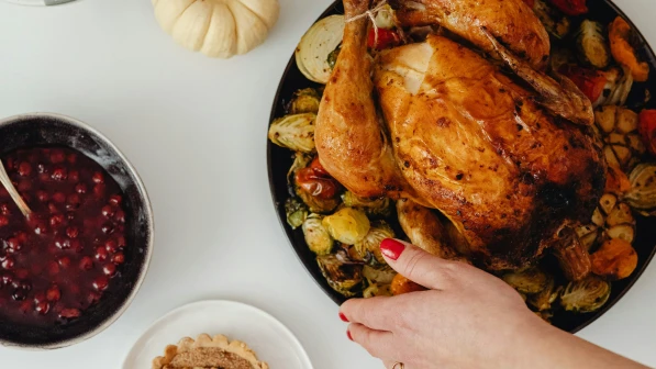 Roasted whole chicken on a black plate surrounded by grilled vegetables, with a bowl of cranberry sauce and a small white pumpkin nearby on a white surface.