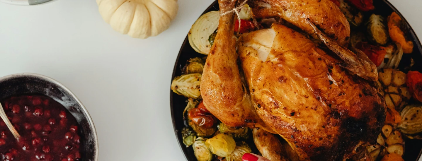 Roasted whole chicken on a black plate surrounded by grilled vegetables, with a bowl of cranberry sauce and a small white pumpkin nearby on a white surface.