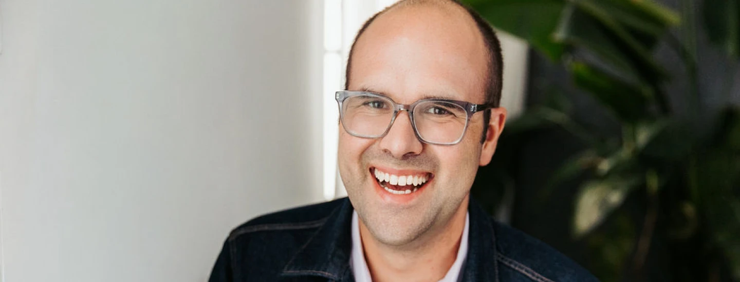 A smiling person wearing glasses, a light-colored shirt, and a dark jacket stands indoors near a plant.