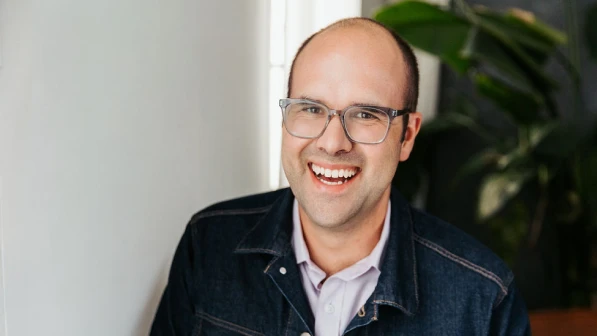 A smiling person wearing glasses, a light-colored shirt, and a dark jacket stands indoors near a plant.
