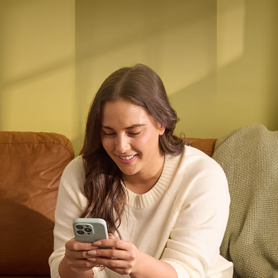 Young woman sitting on a brown couch, smiling while looking at her smartphone.