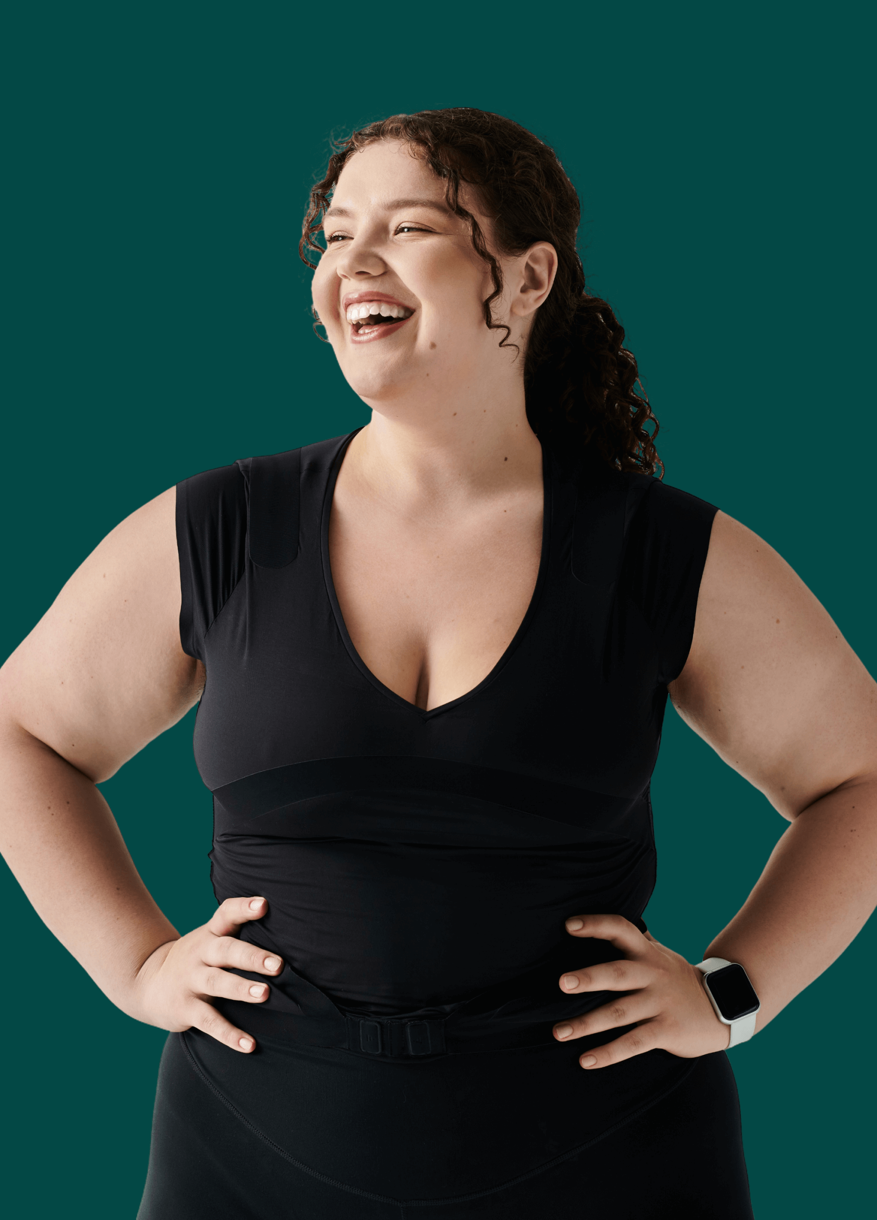 Smiling woman with curly hair in a black top and smartwatch, hands on hips against a solid teal background.