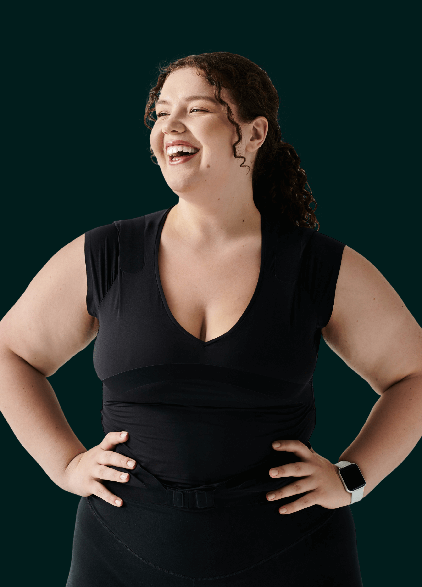 Smiling woman with curly hair wearing a black athletic top and white smartwatch posing with hands on hips against dark background.