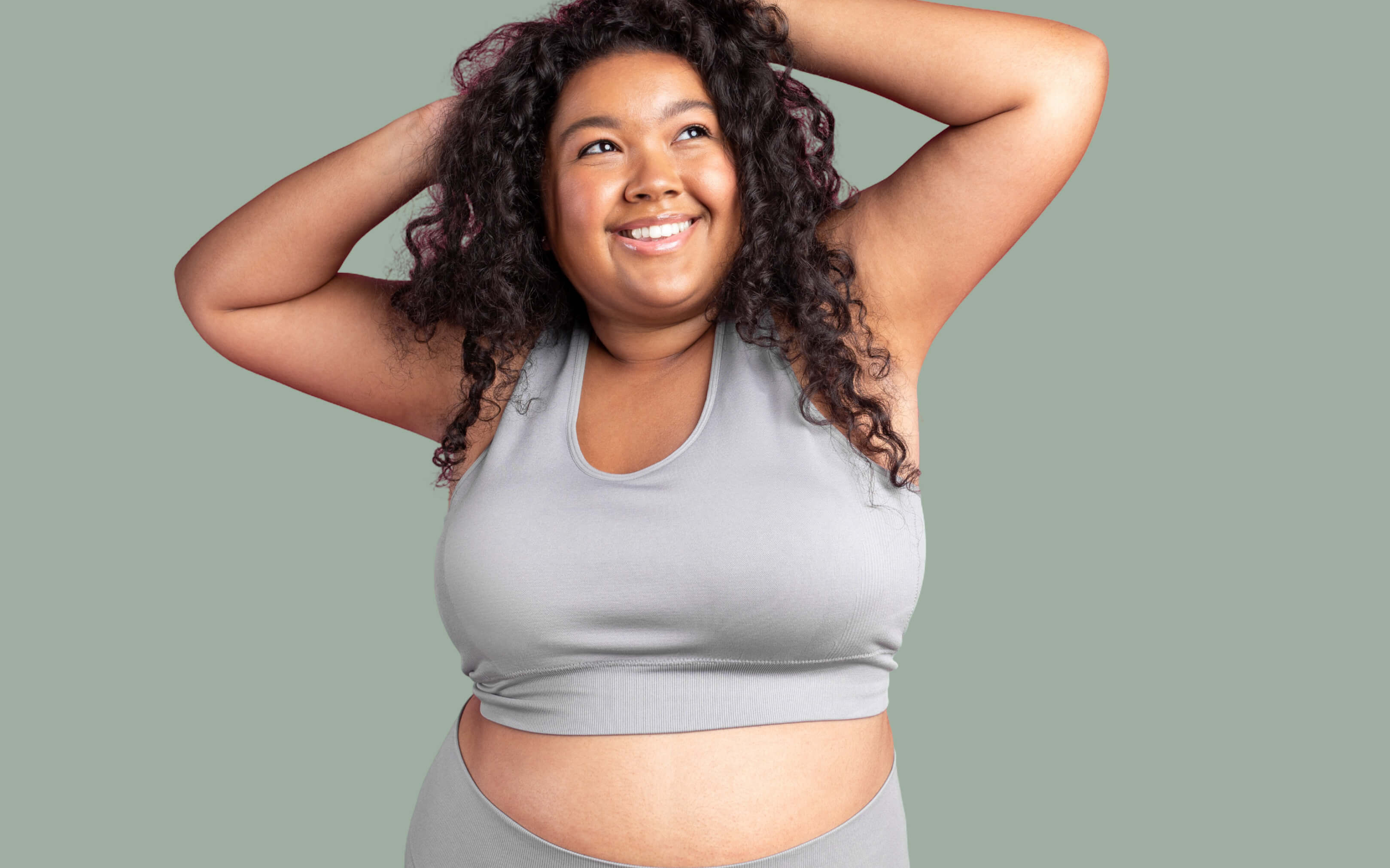 Smiling woman with curly hair in a light gray sports bra lifting her arms behind her head against a solid muted green background.
