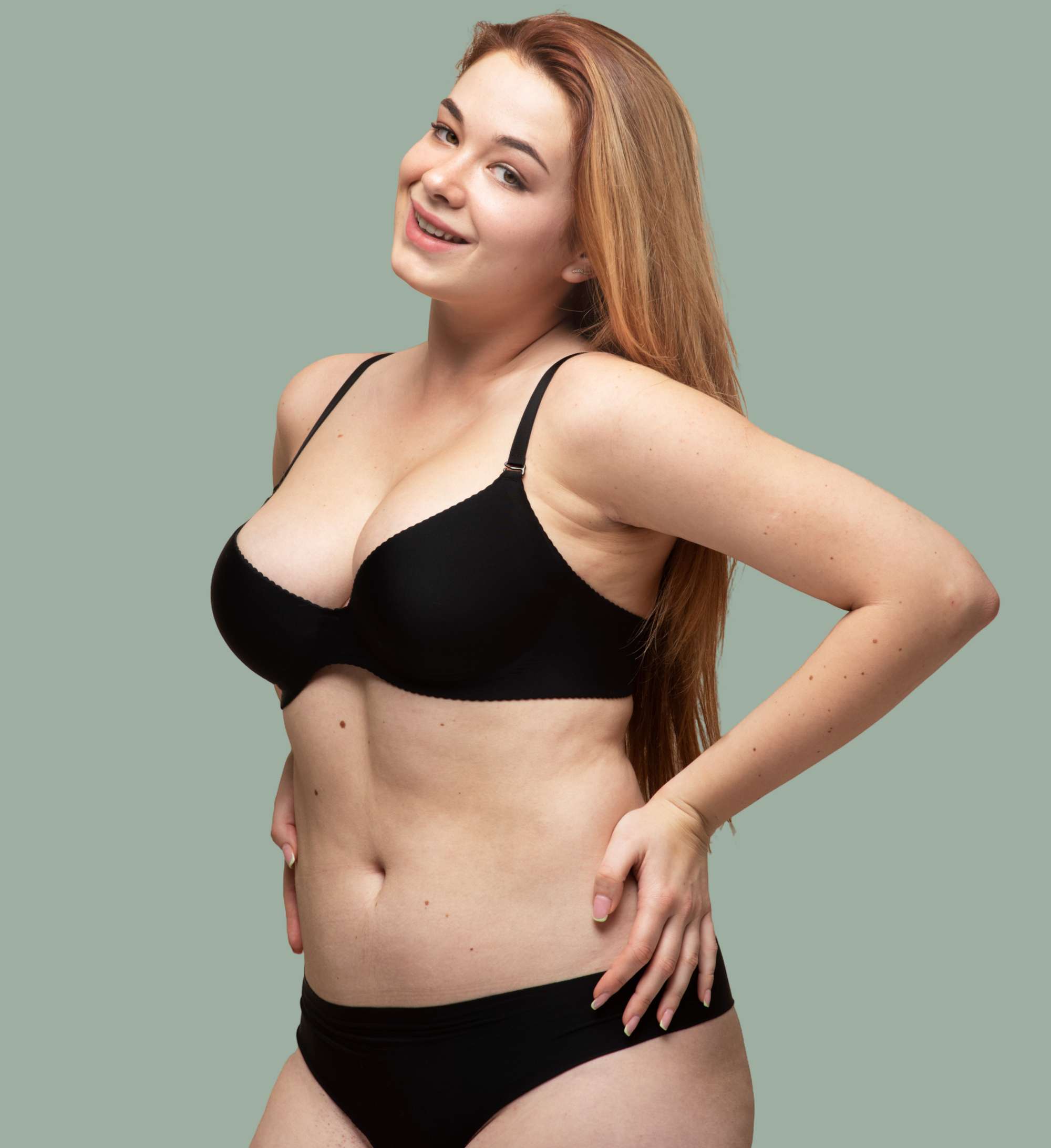 Smiling woman with long light brown hair wearing black bra and underwear against a plain gray-green background.