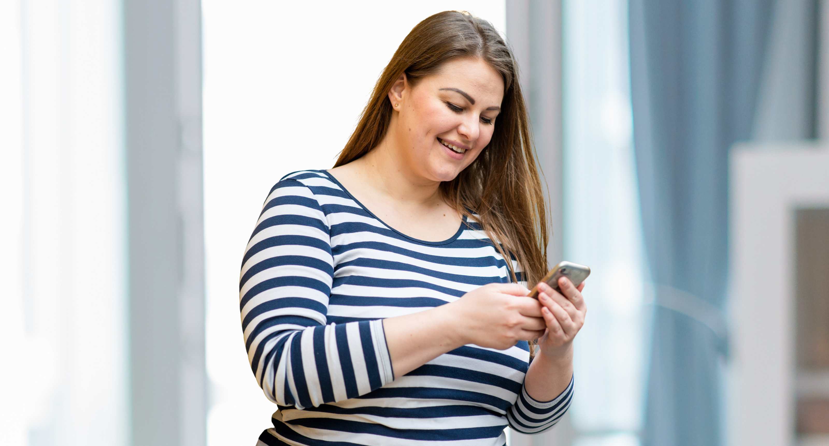 Smiling woman in a blue and white striped shirt looking at her smartphone indoors.