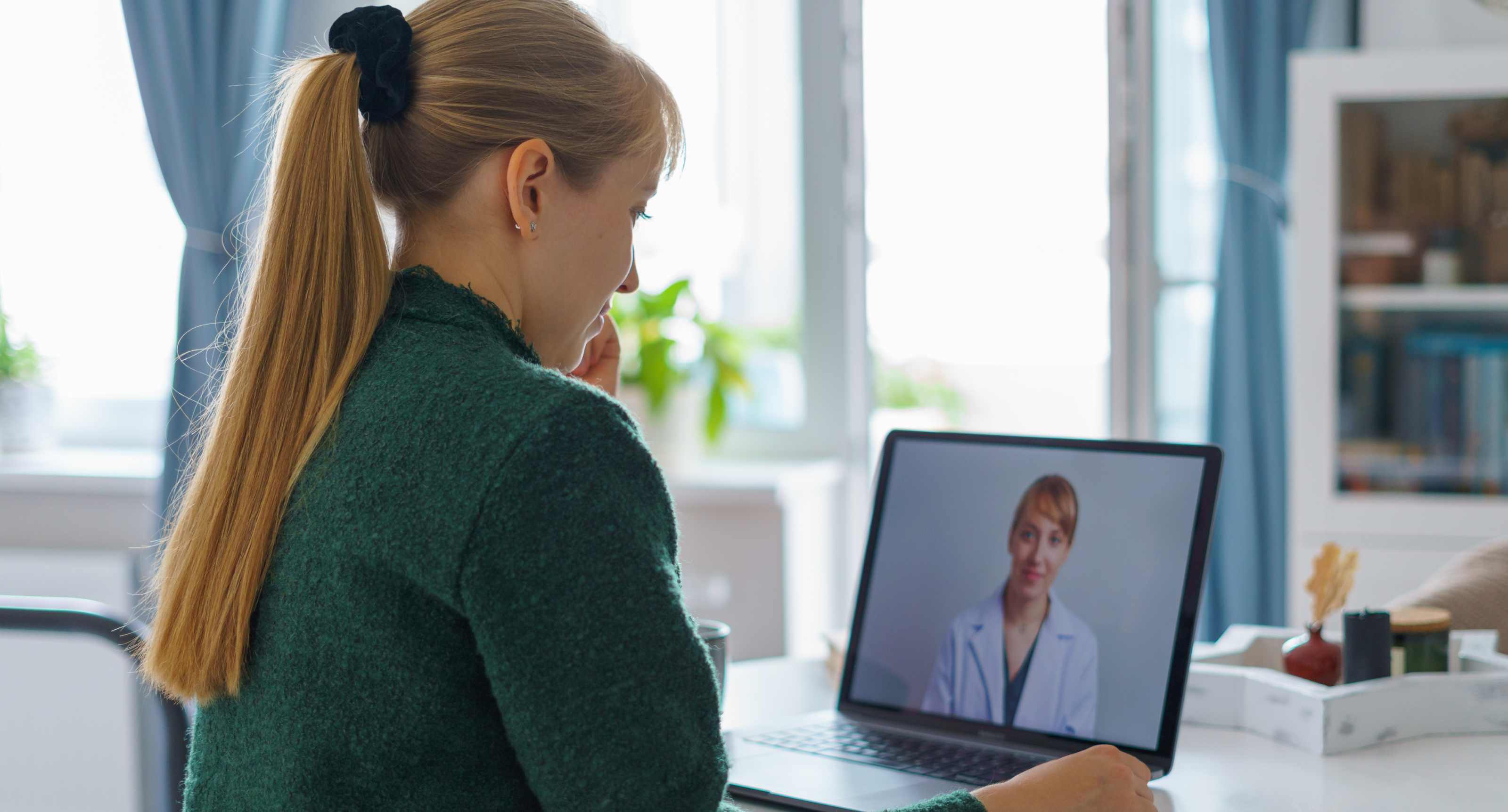 Woman with long blonde hair in a green sweater having a video call with a female doctor on a laptop.