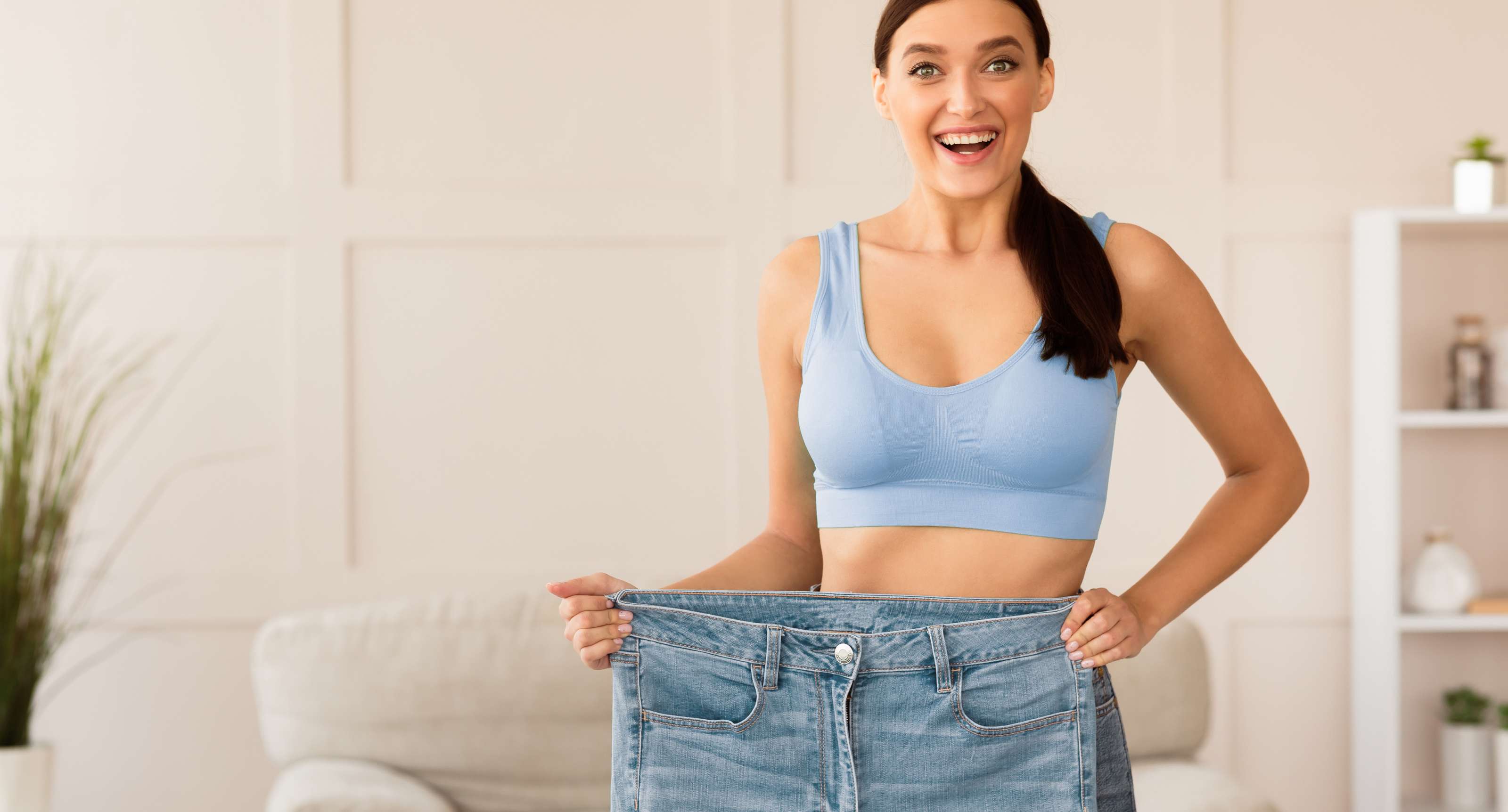 Smiling woman in a blue sports bra holding out the waistband of oversized jeans showing weight loss.
